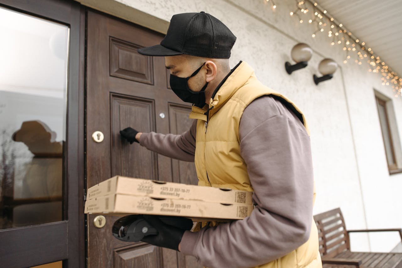 A deliveryman wearing a mask and gloves knocks on a door while holding pizza boxes, emphasizing safety during delivery.